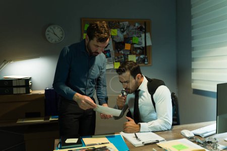 Photo for Two detectives examine paperwork using a magnifying glass in a poorly lit office environment - Royalty Free Image