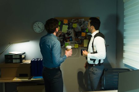 Two male investigators analyzing a disorganized evidence board in a dimly illuminated room, deliberating on a case