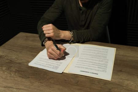 Téléchargez les photos : Criminel en salle d'interrogatoire examine attentivement un accord d'immunité, prêt à signer avec un stylo en main - en image libre de droit