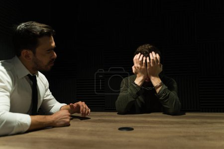 Foto de Abogado hablando con un sospechoso esposado mostrando signos de angustia en una sala de interrogatorios con poca luz - Imagen libre de derechos