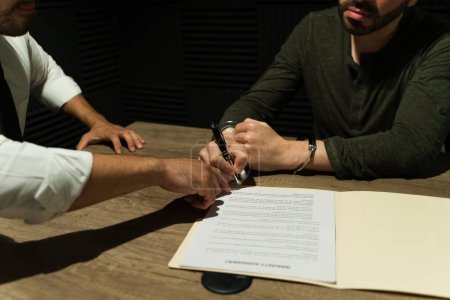 Photo for Cloaked in shadow, a prisoner signs an immunity deal during an interrogation with a detective - Royalty Free Image