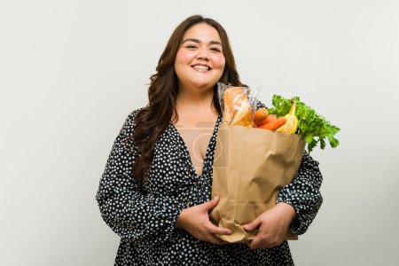 Foto de Retrato de una mujer alegre de tamaño grande posando con una bolsa de papel de productos frescos de granja en un entorno de estudio - Imagen libre de derechos