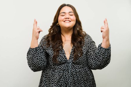 Foto de Mujer esperanzada y optimista de talla grande con los ojos cerrados y los dedos cruzados emitiendo brillo - Imagen libre de derechos