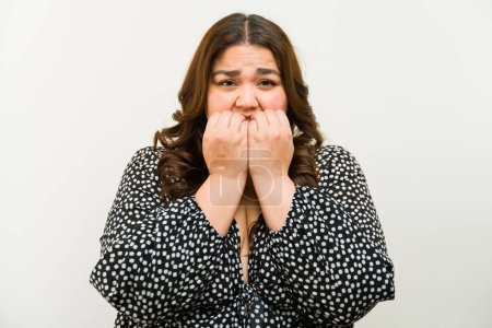 Photo for Nervous plus-size woman nervously biting her nails in a studio against a white background - Royalty Free Image