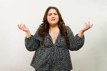 Photo for Expressive plus-size woman shows confusion or doubt with hand gestures against a white studio background - Royalty Free Image