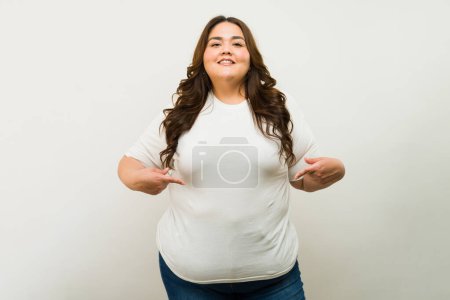Foto de Mujer confiada de talla grande orgullosamente de pie en una camiseta blanca y pantalones vaqueros contra un telón de fondo neutral, sonriendo - Imagen libre de derechos
