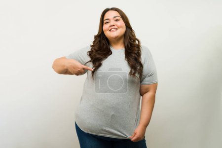 Foto de Linda mujer gorda hispana usando una camiseta gris y apuntándola mientras está de pie sobre un fondo blanco - Imagen libre de derechos