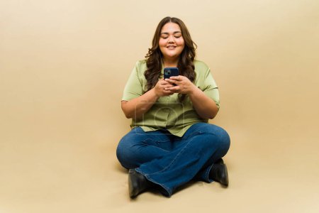 Photo for Happy plus-size woman sitting cross-legged in a studio and using a smartphone - Royalty Free Image