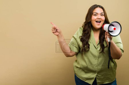 Photo for Confident plus-size woman pointing to copy space and speaking through a megaphone in a studio setting - Royalty Free Image