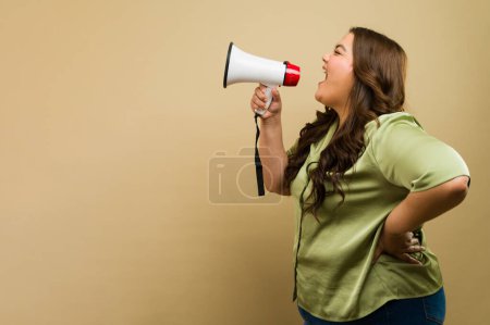 Photo for Cheerful plus-size woman speaking through a megaphone on a beige background in a studio setting - Royalty Free Image