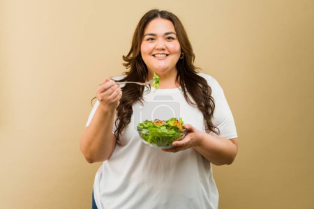 Foto de Mujer curvilínea alegre comiendo un tazón saludable de ensalada sobre un fondo cálido - Imagen libre de derechos