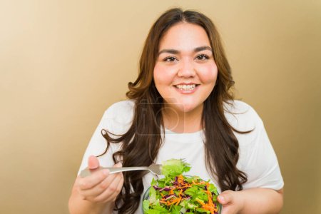 Foto de Mujer alegre y de tamaño grande que promueve la positividad corporal y la alimentación saludable con una ensalada fresca en un entorno de estudio - Imagen libre de derechos