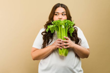 Photo for Serious plus-size woman posing with a bunch of fresh celery in a studio setting and looking towards copy space - Royalty Free Image