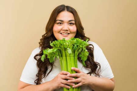 Photo for Cheerful plus-size woman with a radiant smile posing in a studio with a bundle of green celery sticks - Royalty Free Image