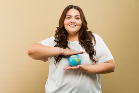 Téléchargez les photos : Femme heureuse de grande taille en tenue décontractée tenant un globe miniature dans un environnement de studio - en image libre de droit