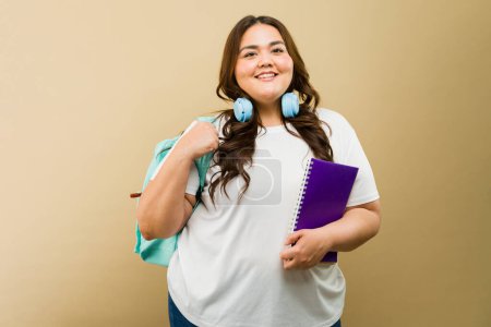 Foto de Alegre mujer de tamaño grande equipada con auriculares y una mochila con un portátil y mirando listo para la escuela - Imagen libre de derechos
