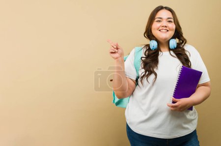 Foto de Estudiante universitaria y mujer de talla grande con ropa casual apuntando a espacio para copiar, preparada para la escuela con un cuaderno - Imagen libre de derechos