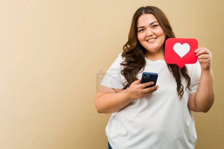Téléchargez les photos : Joyeuse femme de plus de taille tenant une icône du cœur et un téléphone portable dans un cadre studio - en image libre de droit