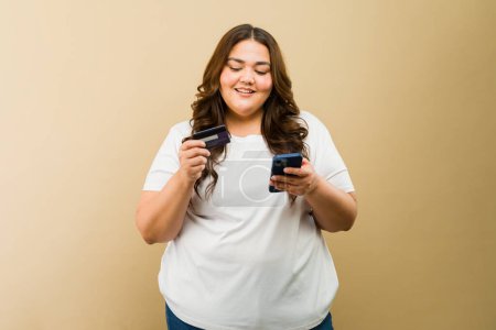 Foto de Atractiva mujer gorda hispana usando su teléfono inteligente y una tarjeta de crédito para hacer algunas compras en línea - Imagen libre de derechos