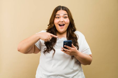 Téléchargez les photos : Heureuse femme latine taille plus pointant son téléphone, soulignant la puissance de la positivité et de la technologie - en image libre de droit