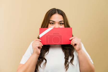 Foto de Retrato de una alegre mujer de talla grande con atuendo casual sosteniendo una tarjeta de regalo roja cerca de su cara en un entorno de estudio - Imagen libre de derechos