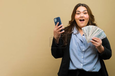 Photo for Happy plus-size woman posing with a phone and a handful of cash in a studio with copy space - Royalty Free Image