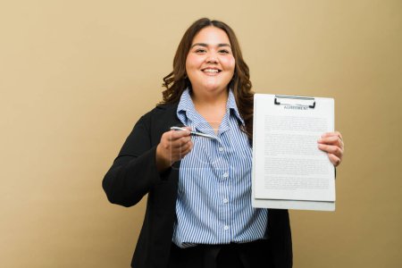 Foto de Mujer sonriente de talla grande sosteniendo un bolígrafo y un contrato, ofreciendo una oportunidad de firma en un estudio - Imagen libre de derechos