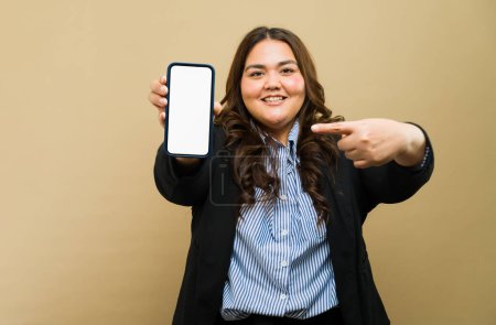 Foto de Retrato de una mujer alegre de talla grande con atuendo profesional mostrando una pantalla de teléfono en blanco en un entorno de estudio - Imagen libre de derechos