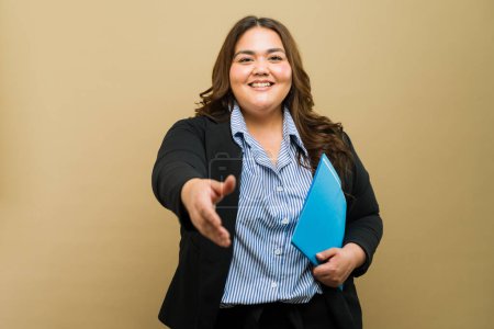 Photo for Plus-size businesswoman in professional attire with a friendly smile, offering a handshake and holding a folder - Royalty Free Image