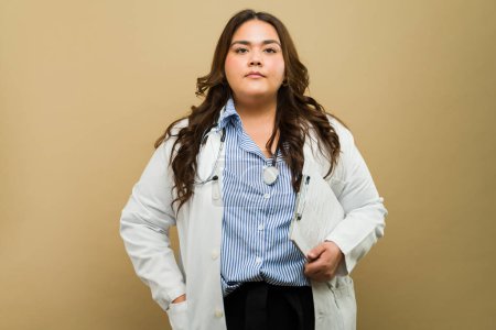 Confident professional female doctor with a stethoscope and lab coat poses in a studio setting, holding a clipboard