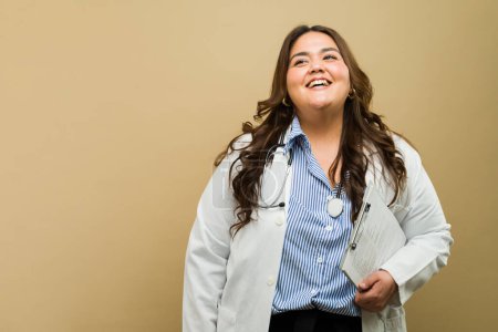 Foto de Mujer feliz de talla grande en ropa médica con portapapeles sobre un fondo beige - Imagen libre de derechos