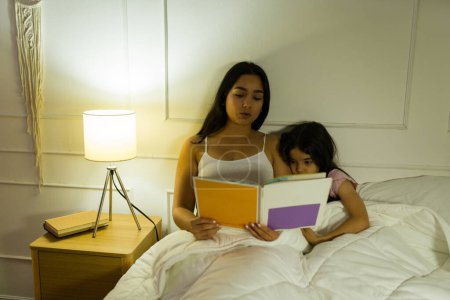 Madre e hija leyendo un libro juntas en un cálido dormitorio por la noche, compartiendo un momento familiar tranquilo e íntimo