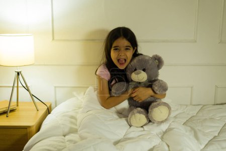 Little girl sitting in her bed at night and hugging her teddy bear while screaming after having a bad dream