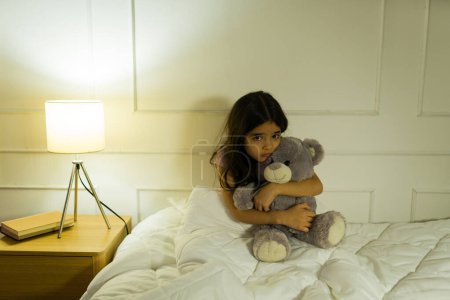 Téléchargez les photos : Petite fille hispanique étreint son ours en peluche étroitement dans la douce lueur de sa lampe de chambre à coucher pendant la nuit et semble affligé de cauchemars - en image libre de droit