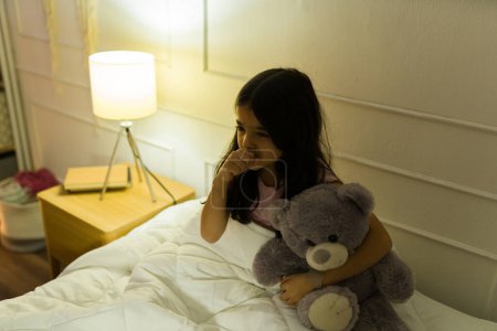 Téléchargez les photos : Petite fille hispanique tenant un ours en peluche dans sa chambre à la maison et toussant et se sentant mal - en image libre de droit