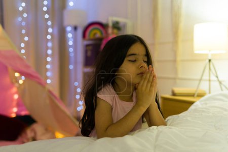 Photo for Young hispanic girl closes her eyes and clasps her hands in prayer in her cozy, warmly lit bedroom at night - Royalty Free Image