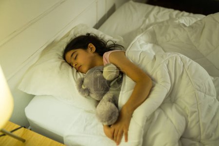 Foto de Chica joven hispana pacífica profundamente dormida, abrazando a un animal de peluche en su cómoda habitación por la noche con una suave iluminación - Imagen libre de derechos