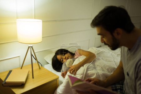 Téléchargez les photos : Père lit une histoire au coucher à sa jeune fille, nichée dans son lit à la maison avec un éclairage chaleureux et confortable illuminant la nuit - en image libre de droit