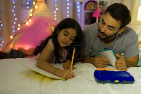 Téléchargez les photos : Le père attentionné aide sa jeune fille avec ses devoirs dans une chambre confortable et chaleureusement éclairée la nuit, créant une atmosphère aimante - en image libre de droit