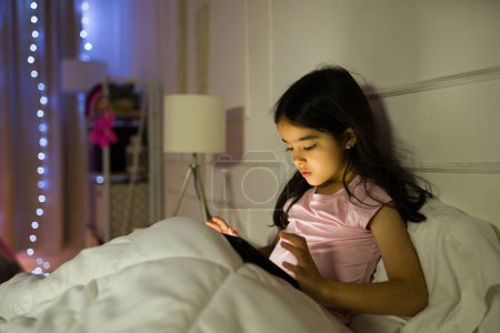 Téléchargez les photos : Petite fille latine utilisant une tablette informatique la nuit et jouant à des jeux vidéo pendant que tout le monde à la maison dort - en image libre de droit