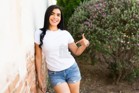 Foto de Joven mujer latina alegre con camiseta blanca lisa y pantalones cortos de mezclilla al aire libre, apuntando al diseño de la camisa, perfecto para maquetas - Imagen libre de derechos