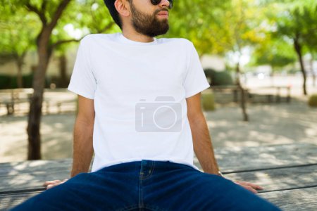 Photo for Closeup of hispanic man wearing a plain white t-shirt, ideal for mockup designs, posing casually in a sunny park setting - Royalty Free Image