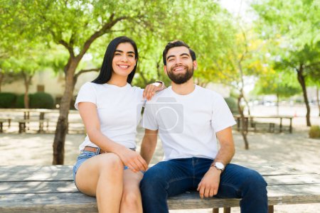 Foto de Alegre pareja hispana joven con camisetas blancas maquetas disfrutan de un día soleado juntos en un parque, mostrando sonrisas brillantes - Imagen libre de derechos