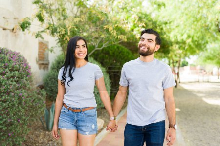 Téléchargez les photos : Jeune couple hispanique souriant tenant la main et marchant ensemble en plein air, portant des t-shirts gris vierges adaptés aux maquettes de marque - en image libre de droit