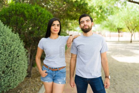 Foto de Hermosa pareja hispana joven posando con confianza en camisetas grises en blanco listas para maquetas de marca, de pie en un exuberante entorno de parque - Imagen libre de derechos