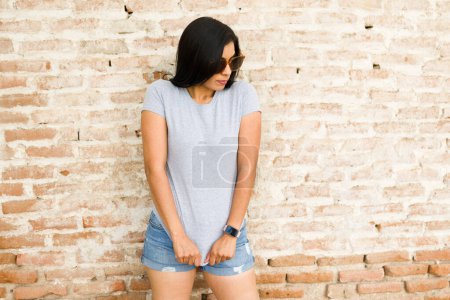 Foto de Mujer hispana joven con una camiseta gris en blanco lista para la marca, de pie sobre un fondo rústico de pared de ladrillo - Imagen libre de derechos