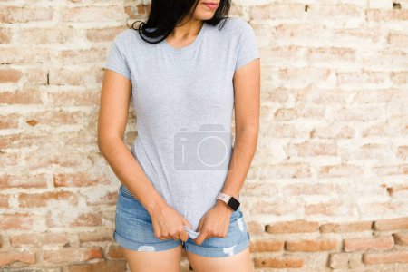 Photo for Hispanic woman wearing a grey t-shirt and blue shorts poses against a brick wall, ideal for fashion mockups and everyday attire - Royalty Free Image