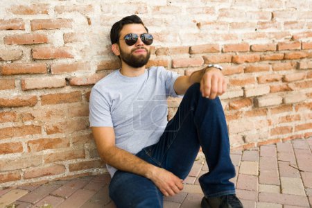 Téléchargez les photos : Élégant jeune homme hispanique portant un t-shirt gris et des lunettes de soleil s'assoit détendu contre un mur de briques, idéal pour les maquettes - en image libre de droit