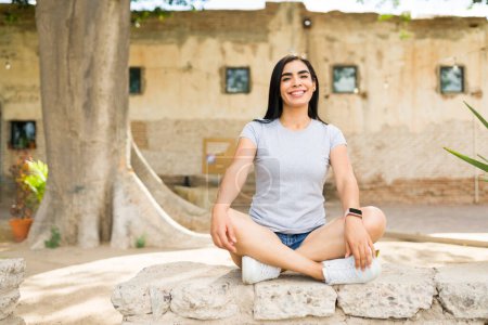 Photo for Cheerful Hispanic woman wearing a blank gray t-shirt sits casually outdoors, perfect for a mockup with a sunny backdrop - Royalty Free Image