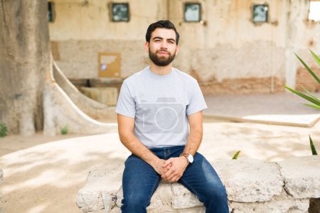 Foto de Sonriente joven hispano con una camiseta gris lisa sentada casualmente al aire libre, perfecta para presentaciones de maquetas de ropa - Imagen libre de derechos